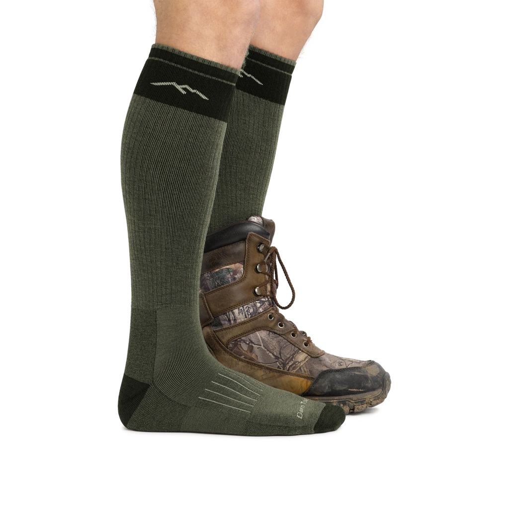 A person wearing olive green knee-high athletic socks with a brown and camouflage hunting boot, shown against a plain white background.